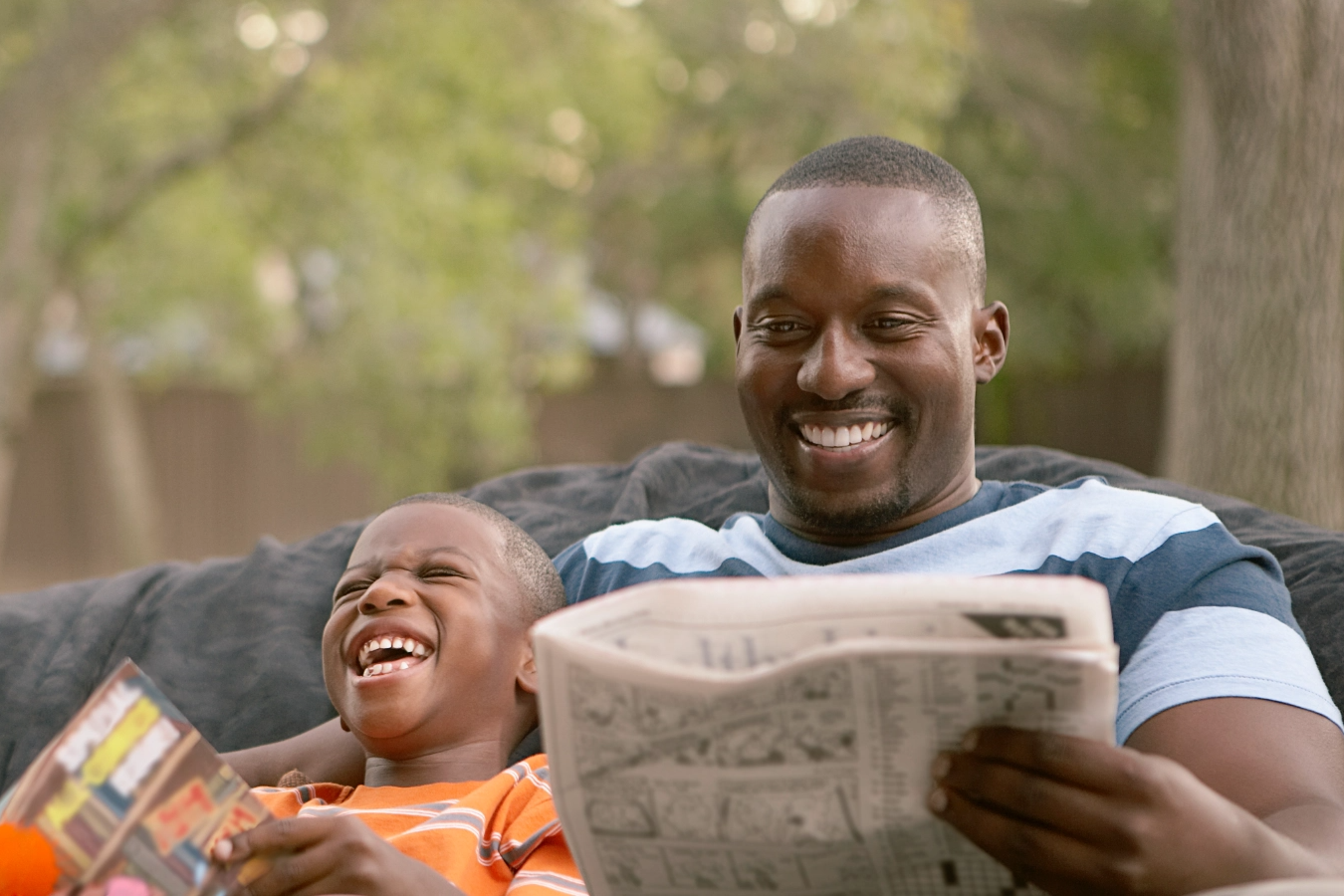 A man, holding a newspaper, and a boy, holding a library book, sit next to each other and laugh.